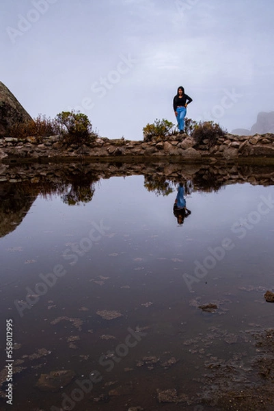 Fototapeta latin woman taking a picture of herself on some rocks that cross a lagoon, where you can see the reflection of the sky and the woman.