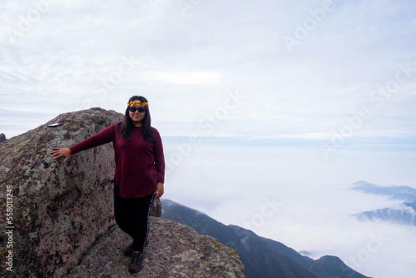 Fototapeta latina woman smiling, standing on the edge of a cliff with a cloud bed in the background