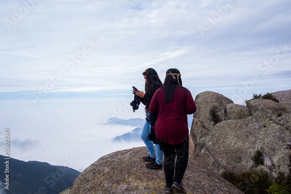 Obraz 2 latina girls, walking to the edge of the cliff to take pictures with their cell phones of the cloud bed.
