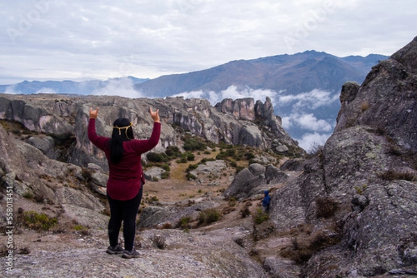 Obraz Latin woman with her back turned and her arms raised, making the sign of the metal, standing on the edge of a rock, looking towards the valley.