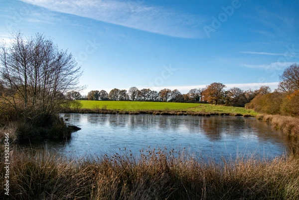 Fototapeta Partly frozen lake with green fields in the Background