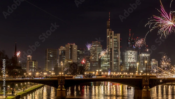 Fototapeta New years eve with fireworks above the skyline of Frankfurt - Main at night at a cold day in winter with colorful reflections in the water.