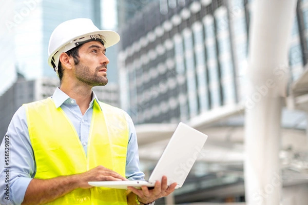 Fototapeta Caucasian man use laptop to working  and wearing a yellow vest and hard hat at the construction site in the city.