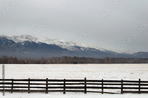 Fototapeta An old wooden fence by the road against the backdrop of mountains and snow.