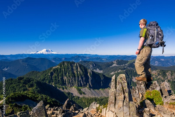 Fototapeta Athletic adventurous male hiker standing on top of a mountain with Mount Rainier in the background during a beautiful sunrise. 
