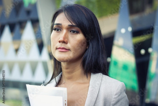 Fototapeta Thai woman portrait with hopeless look holding files folder outside office building. Worried transgender person with documents. Sex discrimination at work, diversity and social inclusion concepts