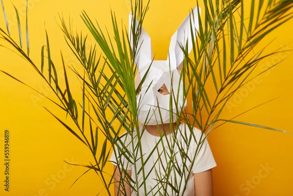 Obraz Indoor shot of anonymous woman wearing white T-shirt and paper rabbit mask standing isolated over yellow background, hiding behind green palm leaves, feels scared.