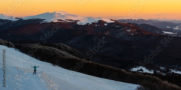 Obraz Backpacker girl admires colorful sunset while standing on top of a snowy mountain during cold winter weather