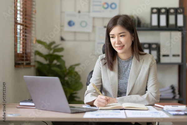 Fototapeta Beautiful Asian businesswoman using her laptop enjoying work, taking notes, reviewing work done.
