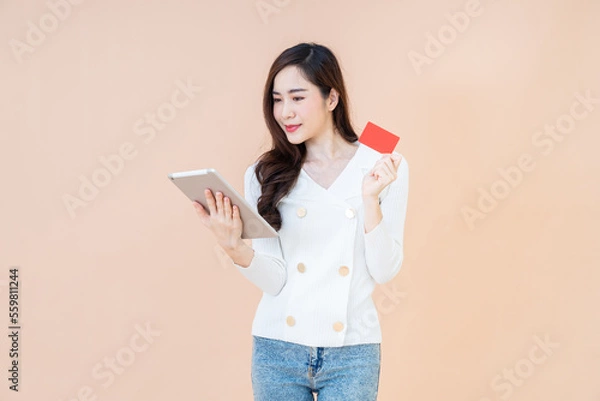 Obraz Portrait of a happy girl showing red credit card while holding mobile tablet isolated over orange background