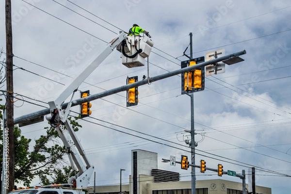 Fototapeta Worker up high in crane bucket repairing traffic light at city intersection on cloudy day with building in background