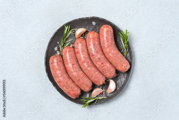 Obraz Raw sausages or bratwurst with with seasonings on black plate over white stone table background. Traditional pork sausages ready to grill. Overhead view