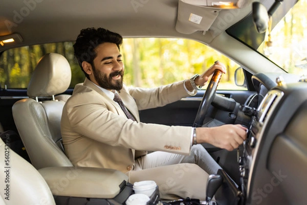 Fototapeta Happy arab young man businessman sitting inside luxury automobile, turning on music on dashboard before driving, handsome arabic man having car trip, side view, copy space