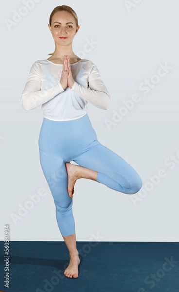 Obraz Young woman in blue and white sport wear keeping hands clasped and one feet on knee, doing tree pose while practicing yoga on mat 