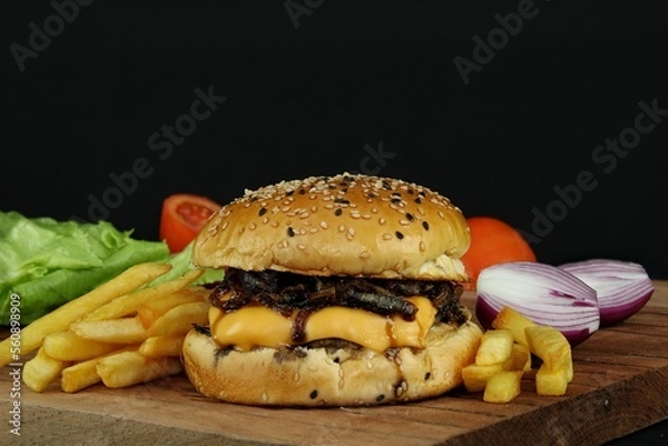 Fototapeta Hamburger with caramelized onion, cheese and french fries, with lettuce, tomato and red onion in the background