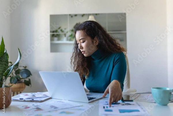 Fototapeta Focused Asian woman using laptop at home, looking at screen, chatting, reading or writing email, sitting on couch, female student doing homework, working on research project online