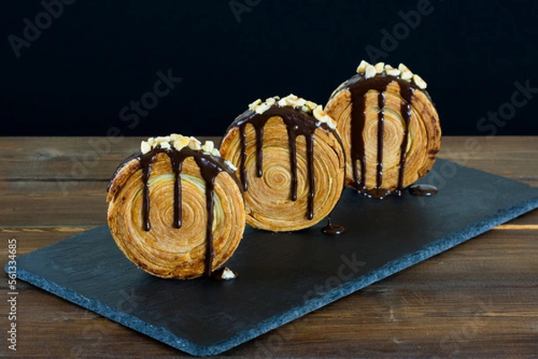 Fototapeta 3 croissants redondos con chocolate y avellanas picadas sobre fuente de pizarra, mesa de madera y fondo negro. New York roll.