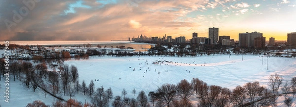 Obraz Wide angle aerial panorama of people sledding down Cricket Hill at sunset with bare trees and snow below and a bright yellow and pink sunset over the Chicago skyline beyond.