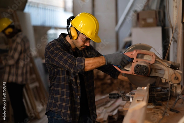 Fototapeta Young carpenter wears safety goggles, earmuffs and yellow helmet, use a circular saw to cut small wood at 45 and 90 degrees for assembly window frame in carpenter shop with coworker, craftsman maker