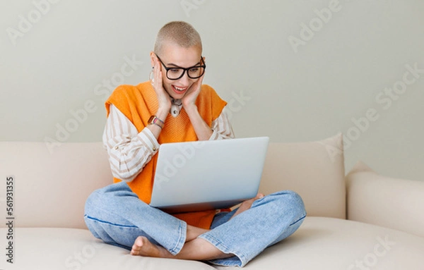 Fototapeta Beautiful woman freelancer with very short hair   working at laptop, makes a video call and happily gestures at home