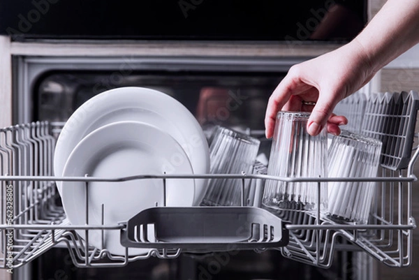 Fototapeta Female hand loading dished, empty out or unloading dishwasher with utensils. Kitchen appliances, lifestyle view. Woman puts a plate in the dishwasher or takes from it. Housewife does her housework 