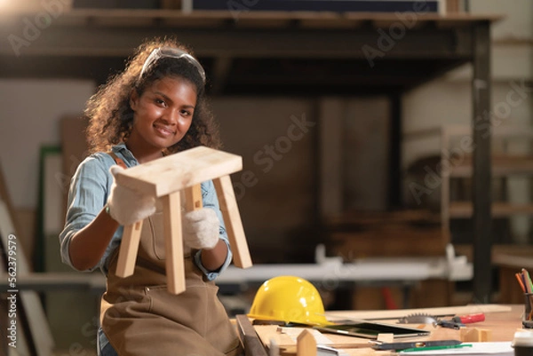 Fototapeta African American young carpenter woman feeling happy and smiling face successfully makes a small chair in hands in a carpentry shop. Professional handmade design furniture on customer order. 