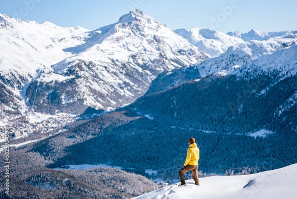 Fototapeta Caucasian man standing on top of a hill looking at winter snow mountain landscape and forests on a sunny day around St. Moritz, Switzerland.
