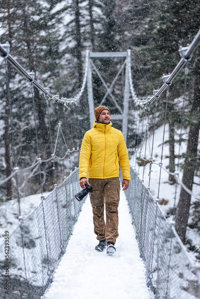 Fototapeta Young caucasian man walking on a hanging bridge on a snowy day holding a camera in winter clothes.