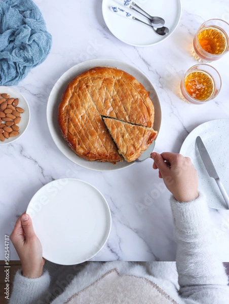 Fototapeta Top view of French galette des rois with cider, hands holding plate and grabbing a piece with handle cake slicer. It's made with puff pastry and creamy almond filling roll in circles shape. 