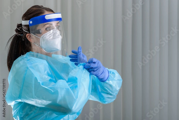 Obraz Nurse is putting on her protective gloves while wearing N95 mask with face shield on her face 