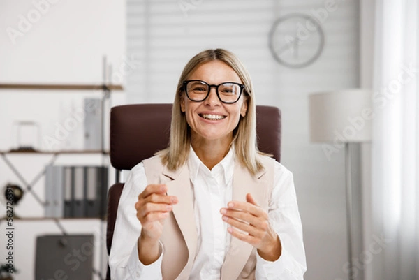 Obraz Smiling mid aged business woman looking at web cam video conference calling in virtual chat meeting, giving remote training webinar class working from home office. Headshot face portrait. Webcam view.