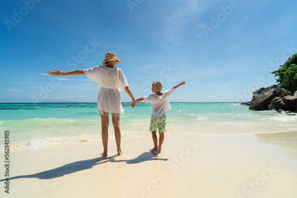 Obraz Tropical photo of mom and son having fun together, holding hands and walking on the sandy beach. Back view. Blue sky.