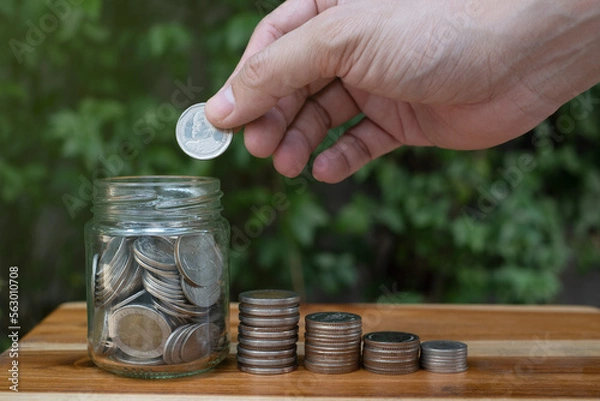 Fototapeta hand of a man saving coin in a jar, and coin on wooden table with green background, symbol of money saving for retirement