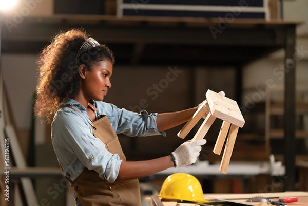 Fototapeta Young black carpenter woman wearing gloves hands, holding small wooden chair in hand after manual assembly. Handmade furniture building based on custom design, crafts joiner maker concept.