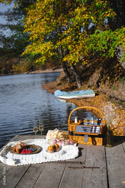 Fototapeta A cozy picnic setup by the lakeside features a woven basket filled with snacks and refreshments. Vibrant autumn leaves surround calm waters, creating a peaceful escape.
