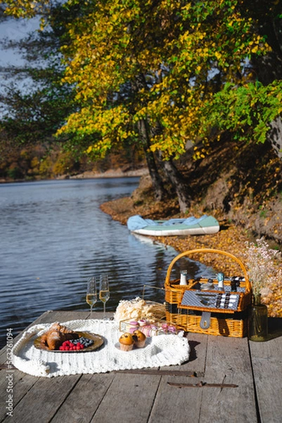 Fototapeta A cozy picnic setup by the lakeside features a woven basket filled with snacks and refreshments. Vibrant autumn leaves surround calm waters, creating a peaceful escape.