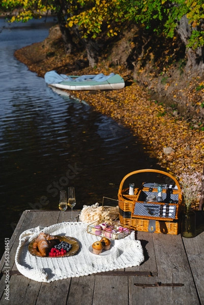 Fototapeta A cozy picnic setup by the lakeside features a woven basket filled with snacks and refreshments. Vibrant autumn leaves surround calm waters, creating a peaceful escape.