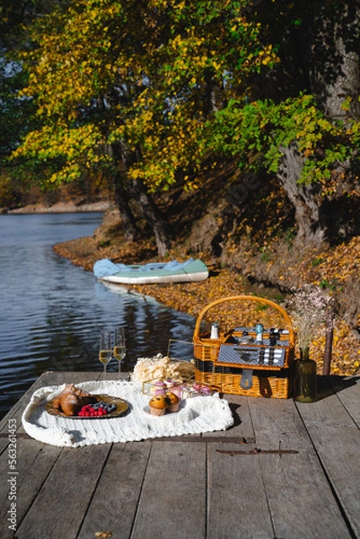 Fototapeta A cozy picnic setup by the lakeside features a woven basket filled with snacks and refreshments. Vibrant autumn leaves surround calm waters, creating a peaceful escape.