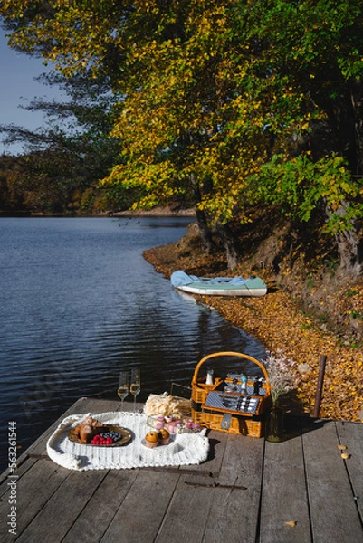 Fototapeta A cozy picnic setup by the lakeside features a woven basket filled with snacks and refreshments. Vibrant autumn leaves surround calm waters, creating a peaceful escape.
