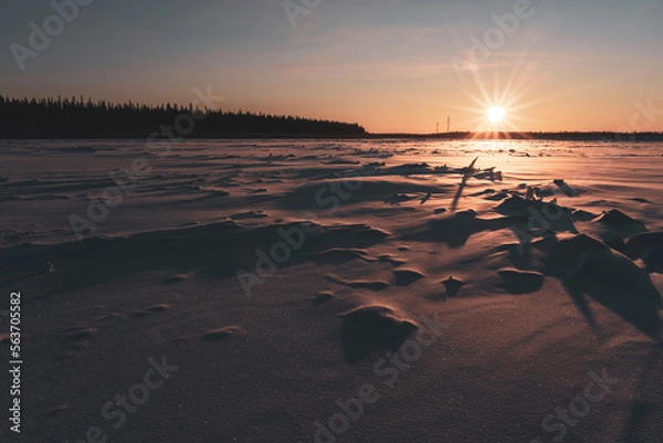 Fototapeta The sun at sunset illuminates the ice on the Vilyui River in Yakutia in winter against the backdrop of a taiga forest with houses in the village of Suntar.