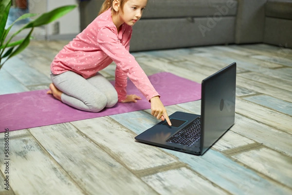 Fototapeta Little girl watching online video on laptop and doing stretching, fitness exercises at home