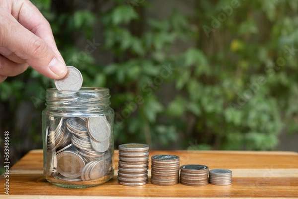 Fototapeta hand of a man saving coin in a jar, with natural lighting and coin on wooden table with green background, symbol of money saving for retirement