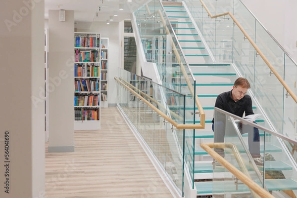 Fototapeta man in library is reading book sitting on steps of glass staircase. education writing scientific work, diploma. thoughtful young adult student or university teacher is learning foreign language