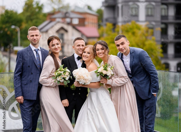 Obraz Front view of happy newlyweds and their friends, wearing in stylish suits and gowns, holding bouquets of flowers , hugging each other and looking at camera during posing on terrace on town background