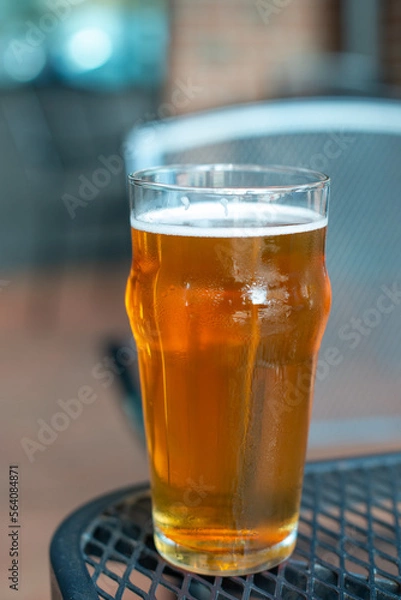 Fototapeta A clear pint drinking glass filled with cold froth from a lager ale. The Irish red ale pint sits on the edge of a metal patio table at a microbrewery. There are tables and chairs in the background.