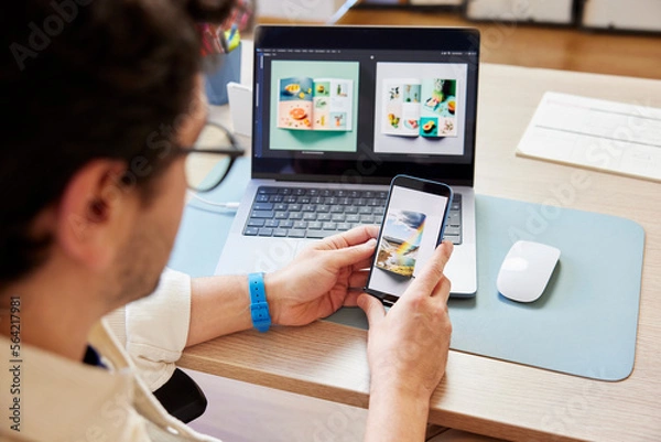 Obraz Crop man using smartphone while working on laptop in studio