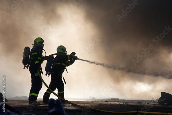 Fototapeta Silhouettes of firemen in smoke, using water to extinguish a massive fire in an industrial building
