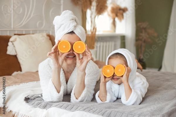 Fototapeta Funny beautiful mother and cute little daughter lying on bed and playing with fresh fruit orange for face mask having spa day toghether at home