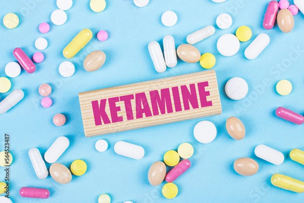 Obraz On a blue background, multi-colored pills and a wooden block with the text KETAMINE. View from above. Medical concept