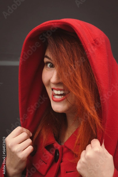 Obraz Close up joyful lady wearing red coat with hood portrait picture. Closeup side view photography with dark grey background. High quality photo for ads, travel blog, magazine, article